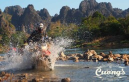 Off-road motorcyclist rides through a shallow river, sending a splash with rocky banks and distant mountains; Cuong's logo in the lower-right corner.
