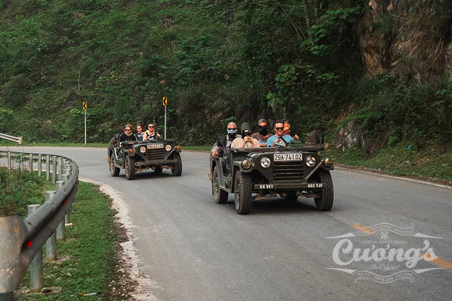 Group of people in open-top vintage military jeeps driving along a winding mountain road surrounded by trees