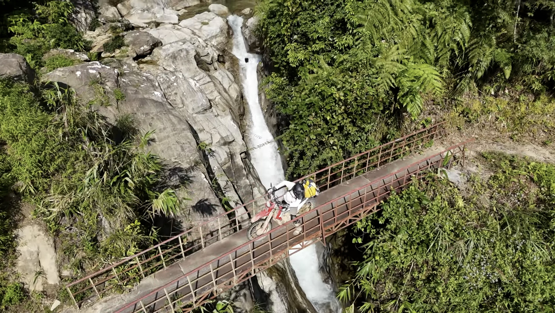 Rider on a dirt bike crossing a rust-colored metal bridge above a narrow waterfall in dense jungle greenery